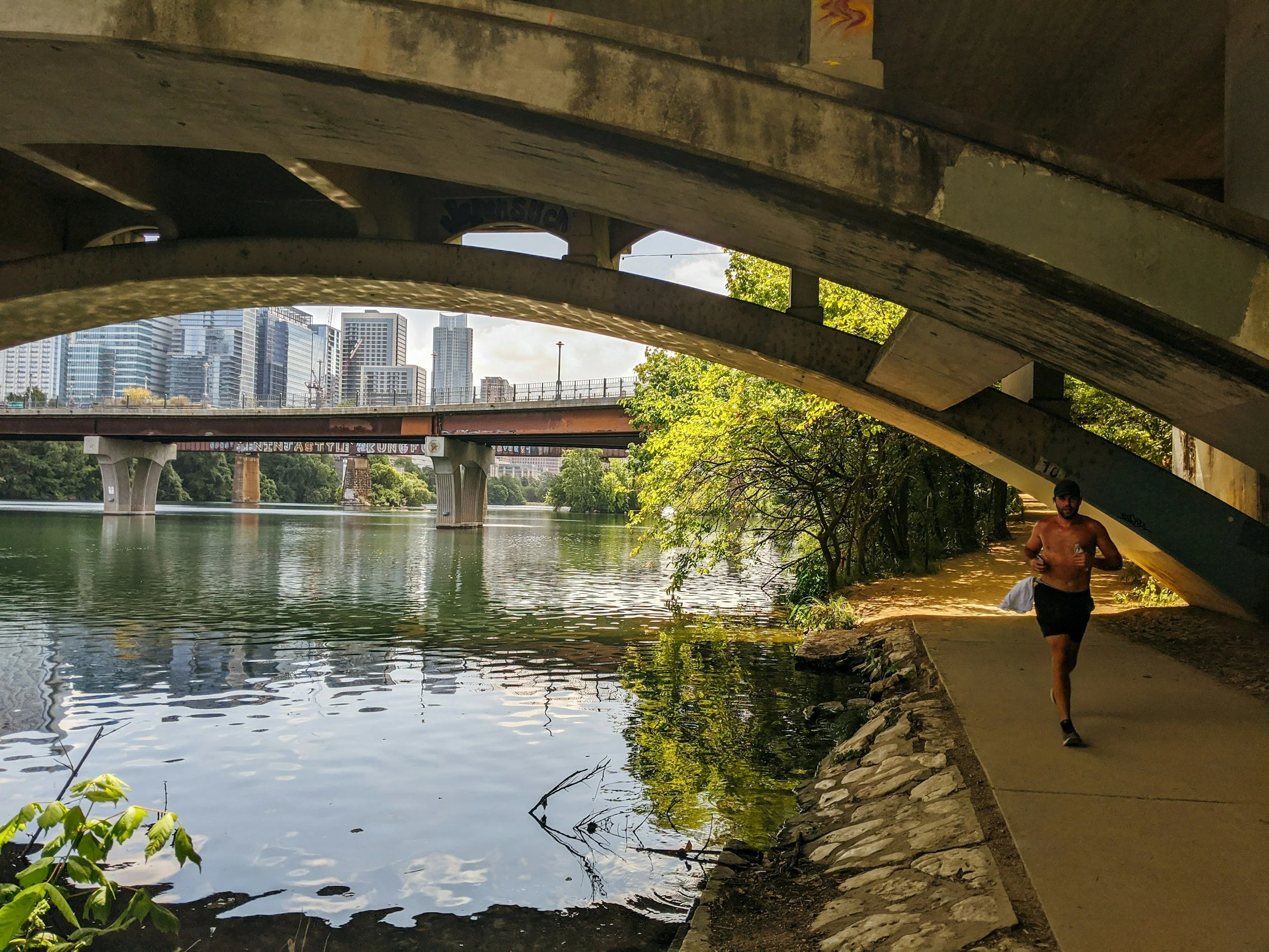 A runner on the Lady Bird Lake trail beneath a bridge, downtown Austin in the distance
